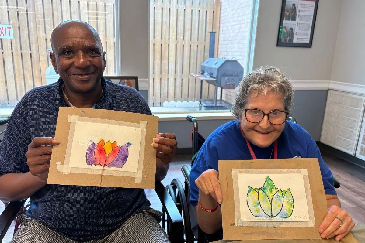 A man and a woman in wheelchairs each hold a watercolor lotus painting.
