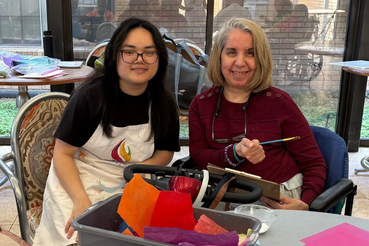 A teaching artist and a seated resident sit at a table with tissue paper collage supplies.