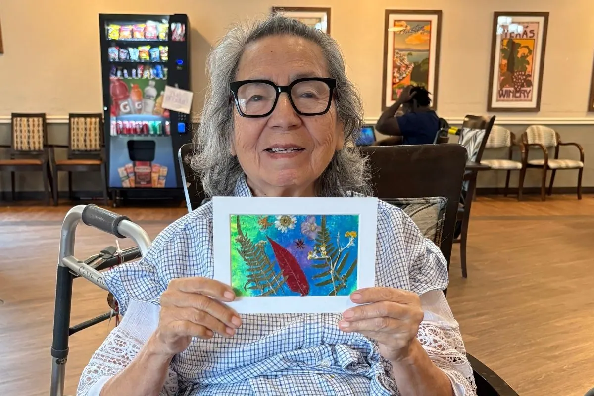 A resident with a walker holds a framed pressed flower and fern artwork.