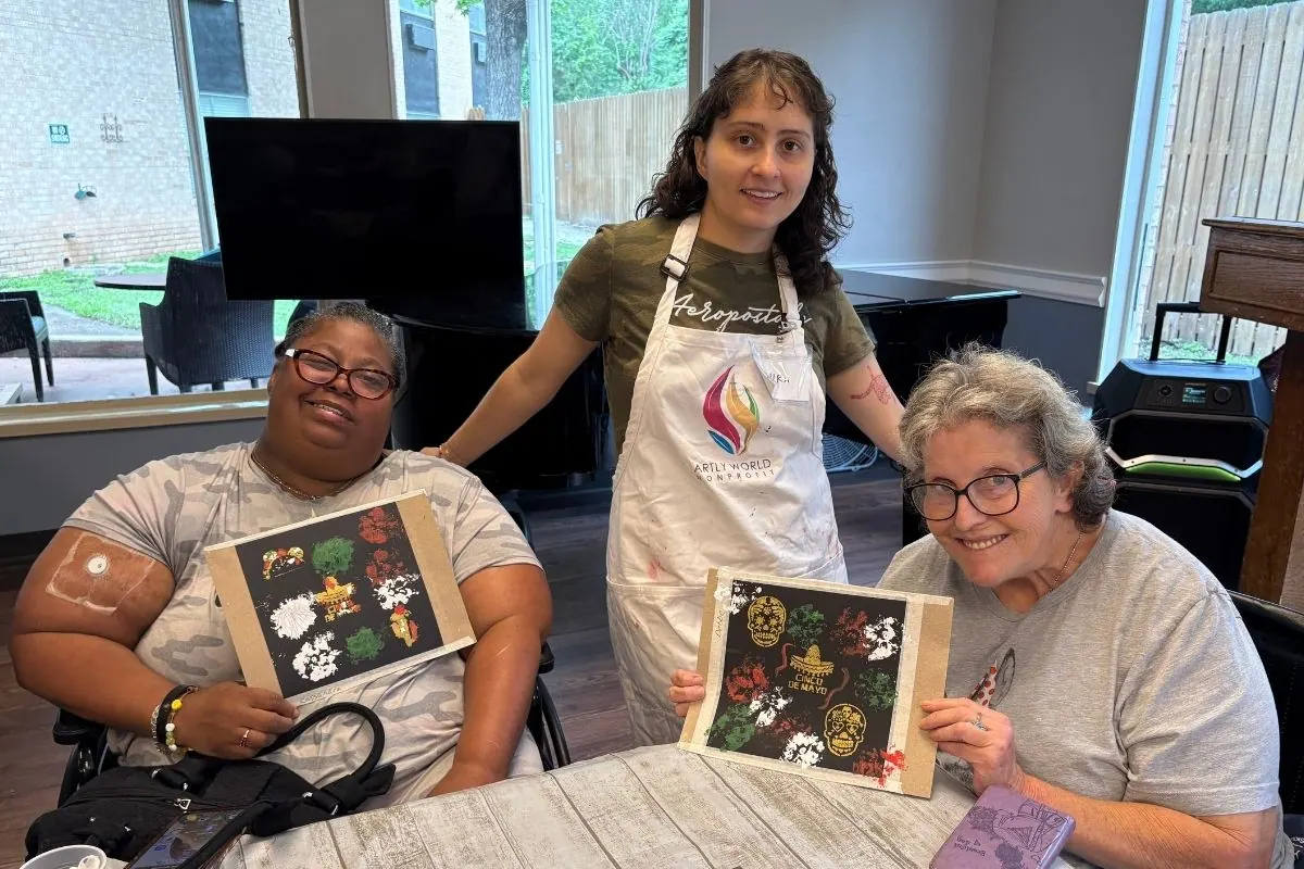 A teaching artist stands between two residents holding paper artwork with cut flowers and skulls.