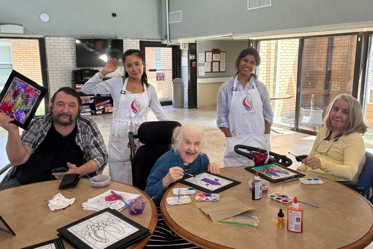 Two teaching artists and three residents paint stained glass style artwork at two round tables.