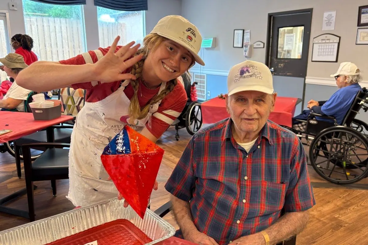 A teaching artist shows a seated resident a red and blue paper diamond craft.