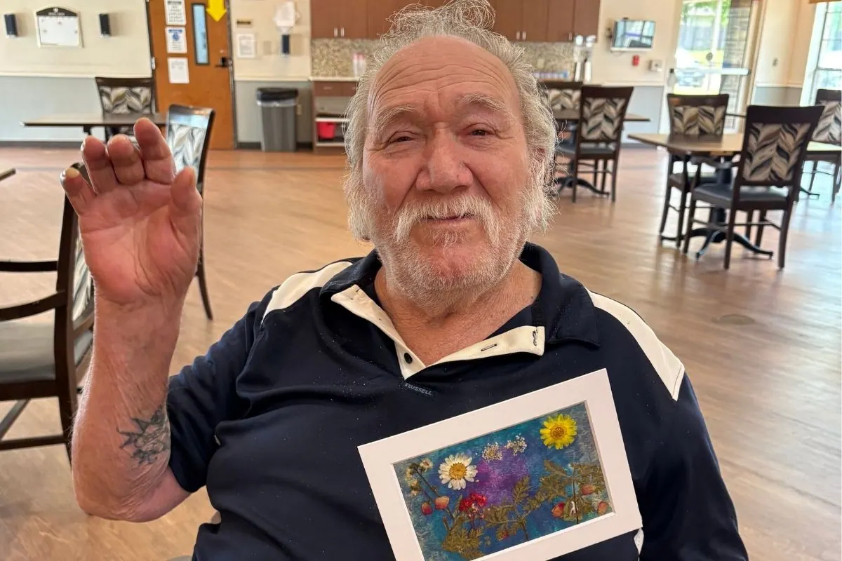 An older man waves while holding a framed pressed flower artwork against his chest.