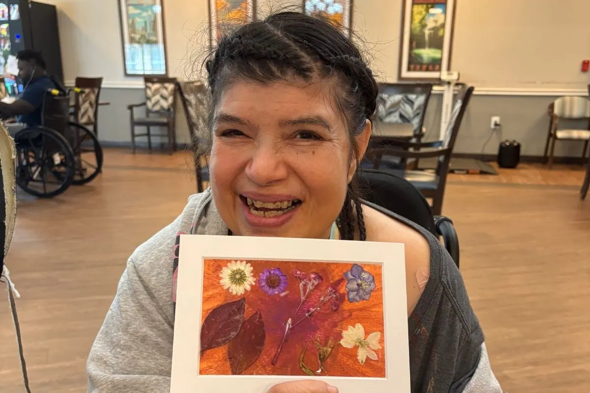 A resident smiles while holding a framed pressed flower artwork on an orange background.