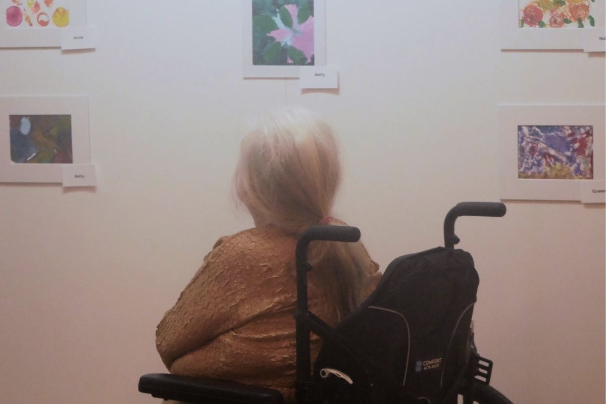 A woman in a wheelchair sits by herself in front of a gallery wall, looking at small framed paintings hung at eye level, each one with a card beside it naming the artist.