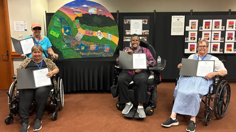 Senior artists hold their certificates in front of a large round landscape painting.