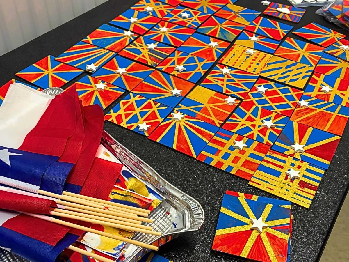 Dozens of hand painted tiles in Texas flag red, white and blue, laid out edge to edge in starburst patterns, with folded Texas flags resting alongside them.