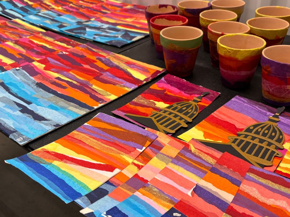 Torn paper collage sunsets in bands of red, orange and violet laid out across a table, several of them cut with a black silhouette of the Capitol dome, with hand painted cups behind.