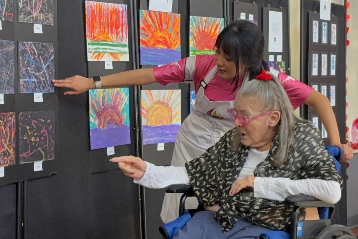 A volunteer beside a seated artist in a wheelchair, both of them reaching toward a wall of framed sunset drawings at the exhibition.