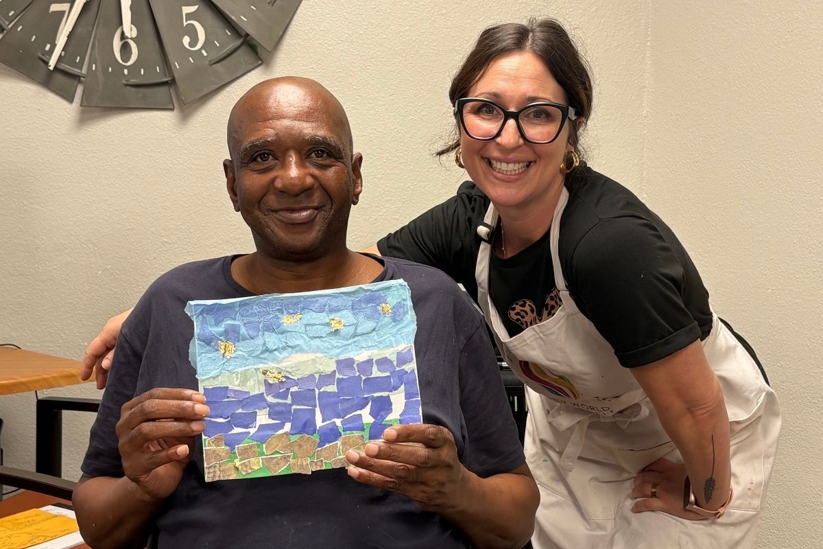 A senior artist holding up the painting he just finished while a volunteer in an apron leans in beside him, both of them smiling, at a weekly studio session.