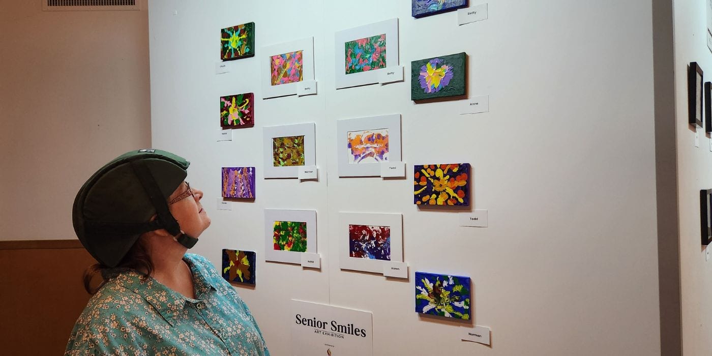 An older artist looks up at a gallery wall of small painted canvases at the Senior Smiles art exhibition, each one with a name card beside it.