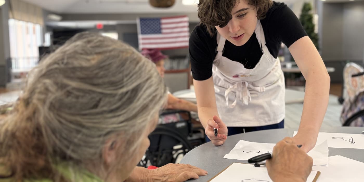 A teaching artist leans across a shared table to help two older adults draw, markers and sheets of paper between them.
